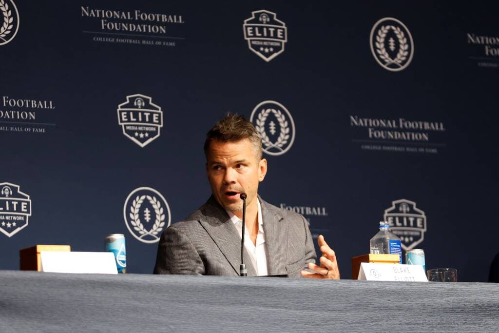 A man in a gray suit speaks at a panel, seated at a table with a microphone, water bottle, and nameplate. Behind him is a dark blue backdrop with National Football Foundation and Elite logo branding.
