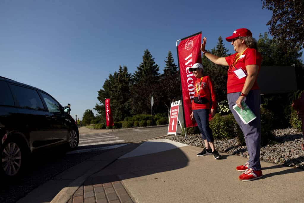 Two people wearing red shirts and hats stand on a sidewalk waving to a black vehicle. Red "Welcome" banners are in the background, and trees line the street on a sunny day.