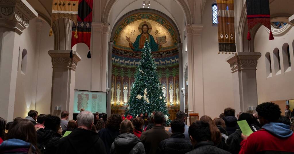 A large group of people stands inside a church, facing a tall decorated Christmas tree in front of a mosaic of Jesus above the altar. Banners and stained glass windows adorn the space.
