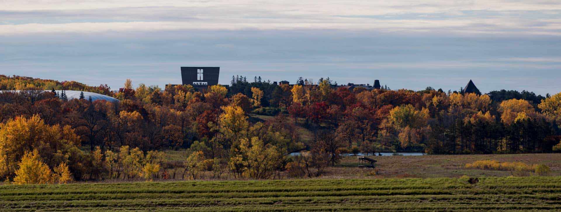 A landscape view of an open field with trees in autumn colors, featuring a modern black building with “h” and “MITTM” on it, and other dark-roofed structures among the trees under a cloudy sky.