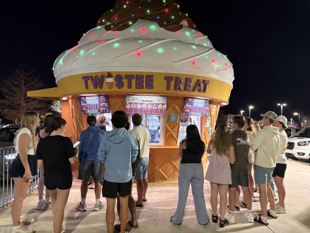 A group of people stand in line at night outside a brightly lit Twistee Treat ice cream stand shaped like a giant ice cream cone, waiting to order.