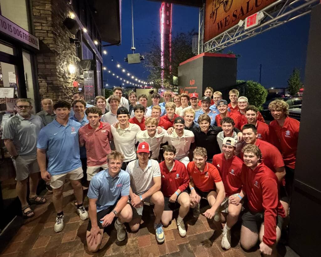 A large group of young men pose and smile together outside a restaurant at night, wearing mostly red, white, and blue shirts. Festive string lights and a neon sign decorate the background.
