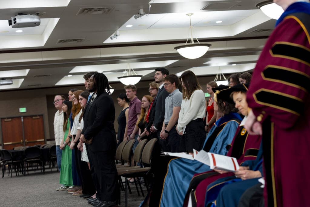 A group of people stands in a line between rows of chairs at an indoor ceremony, while faculty members in academic regalia sit in the foreground reading programs.