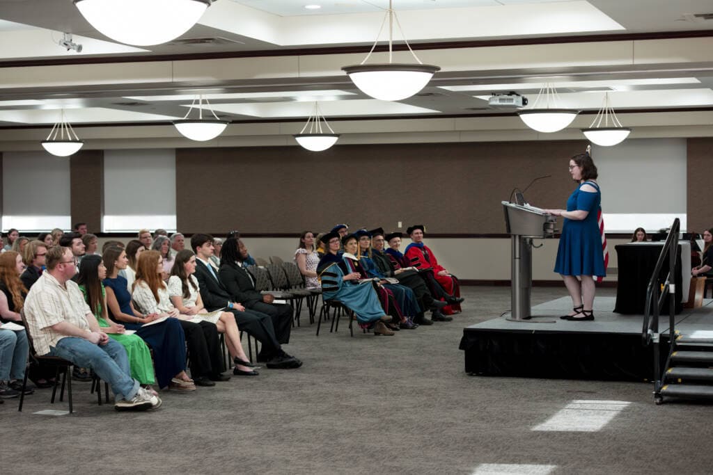 A woman in a blue dress speaks at a podium on a stage in front of an audience, including people in academic regalia and others in casual or semi-formal attire, in a well-lit conference room.