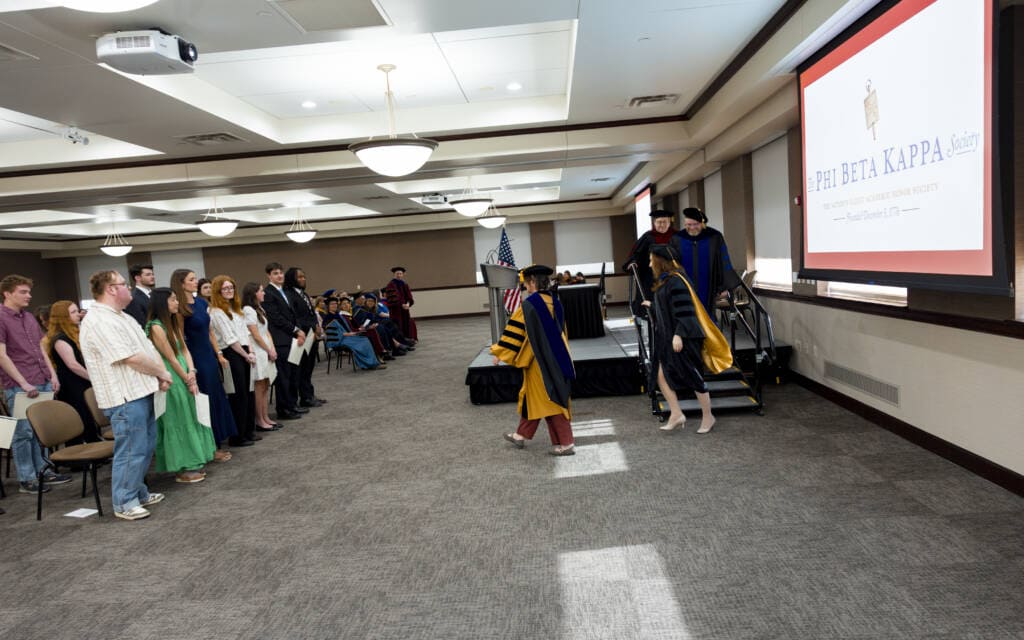 A graduate in a cap and gown walks across a stage during a Phi Beta Kappa ceremony, shaking hands with faculty as an audience of seated and standing people watch in a well-lit conference room.