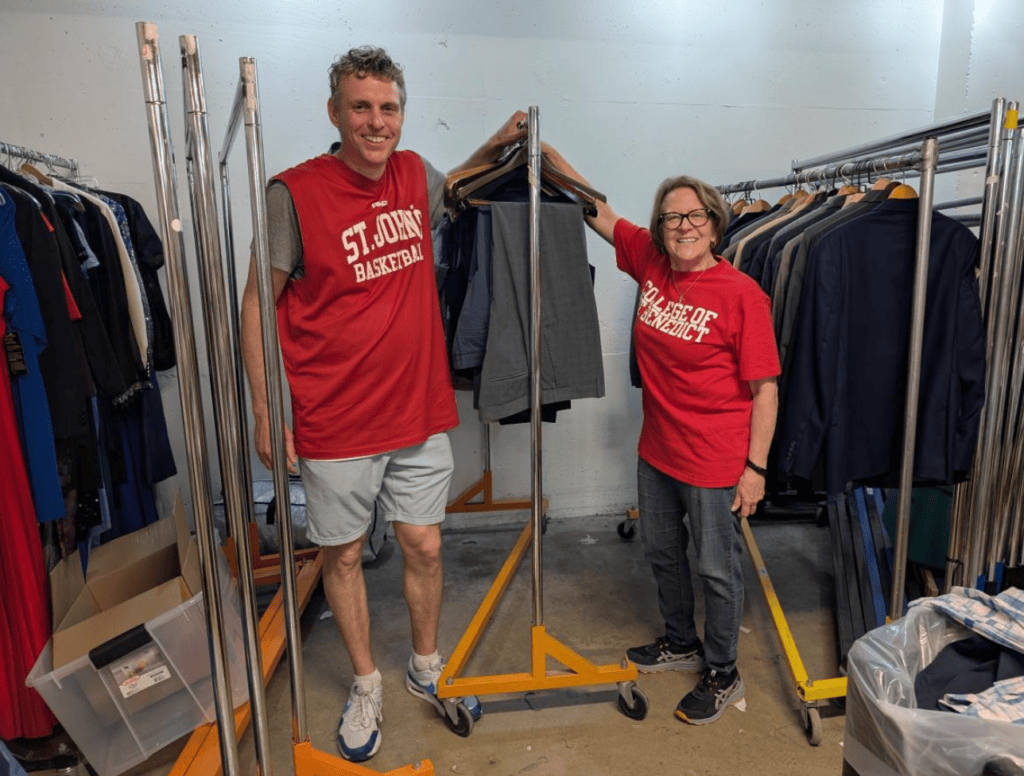 Two smiling adults in red shirts stand next to a clothing rack with suits in a room filled with clothes on hangers and racks. One shirt reads "ST. JOHN'S BASKETBALL," and the other says "A PIECE OF.
