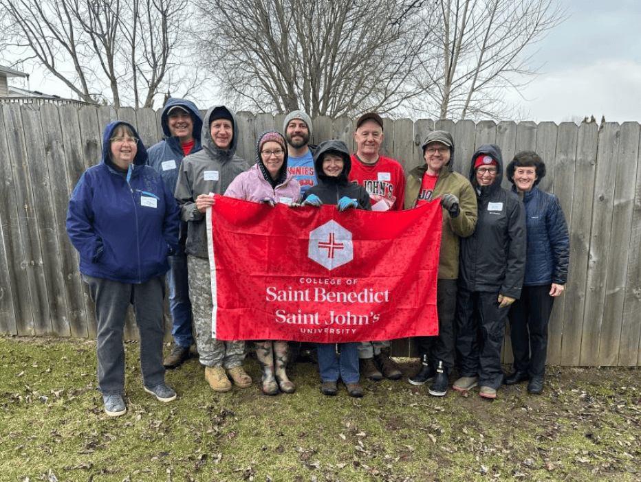 A group of ten people stand outside in front of a wooden fence, holding a red "College of Saint Benedict Saint John's University" banner. They are dressed warmly and smiling, with cloudy skies and bare trees behind them.