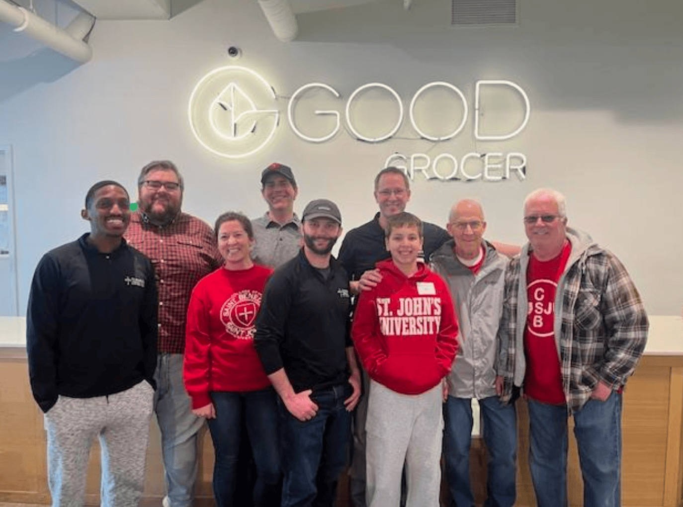A group of ten people, some wearing red St. John’s University clothing, stand and smile together in front of a wall with a “Good Grocer” sign.