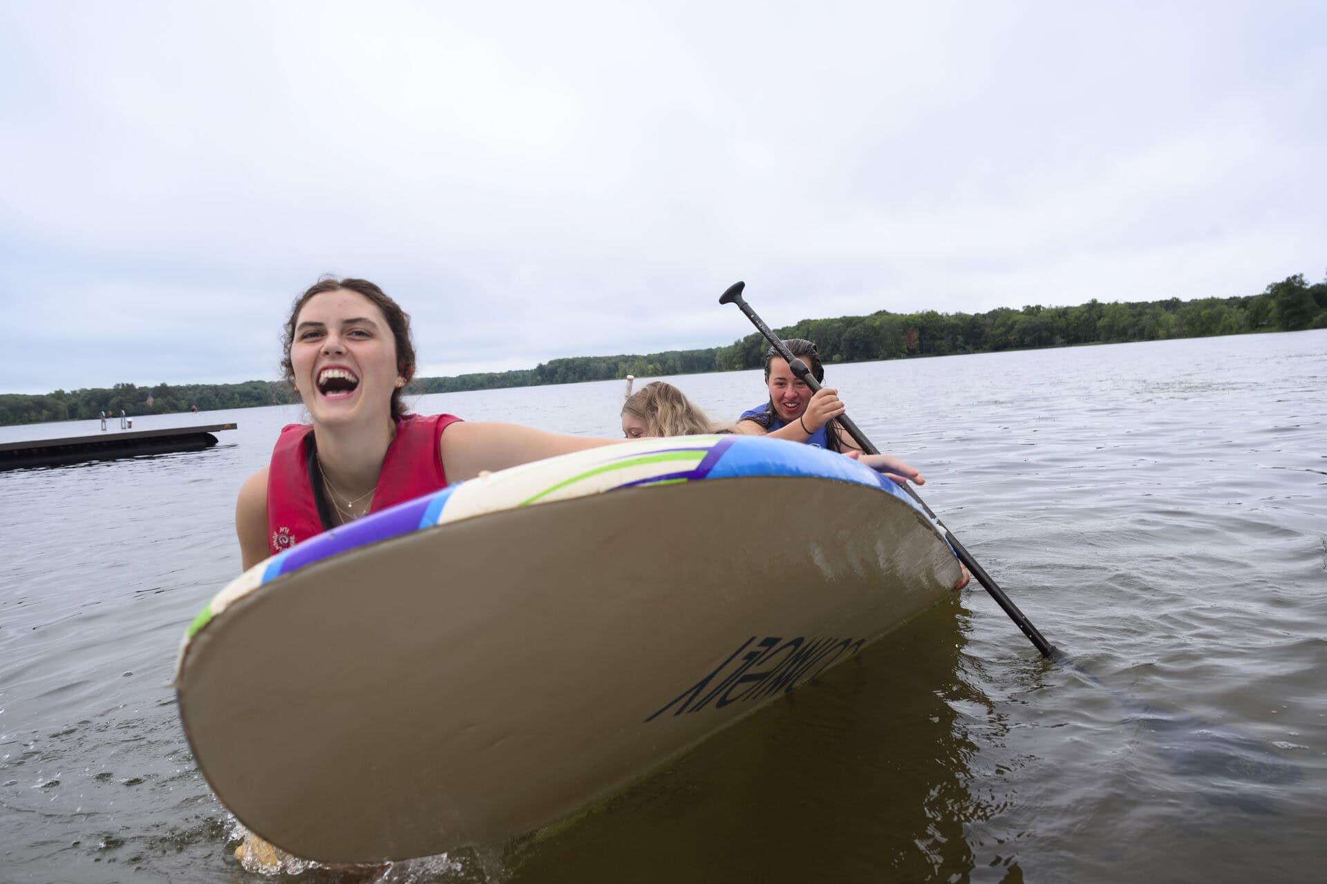Three people on a paddleboard in a lake, smiling and laughing as they approach the shore, with trees and a dock visible in the background on a cloudy day.