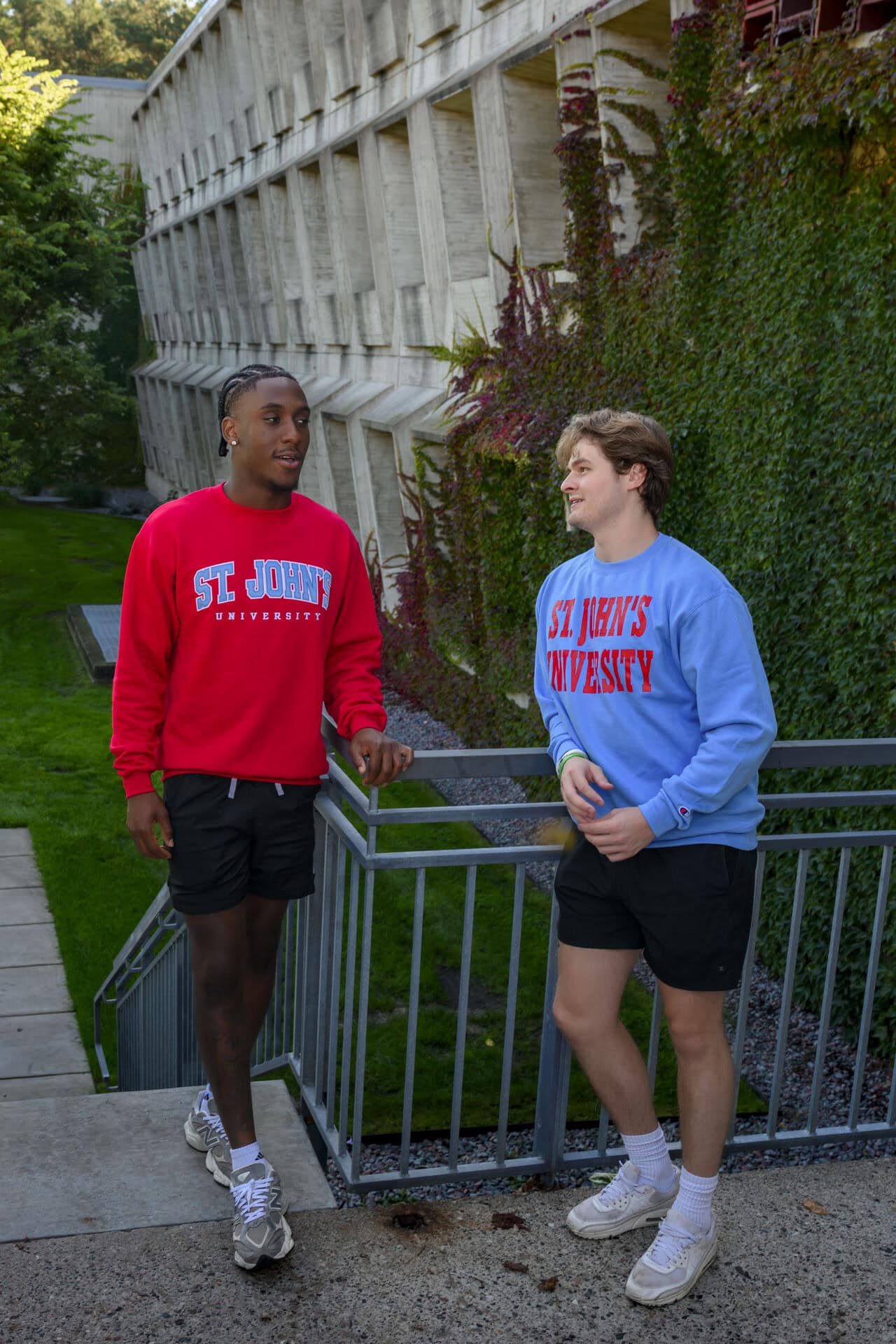 Two young men wearing "St. John's University" sweatshirts and shorts stand outside a modern building, chatting and smiling near a metal railing and lush ivy-covered wall.