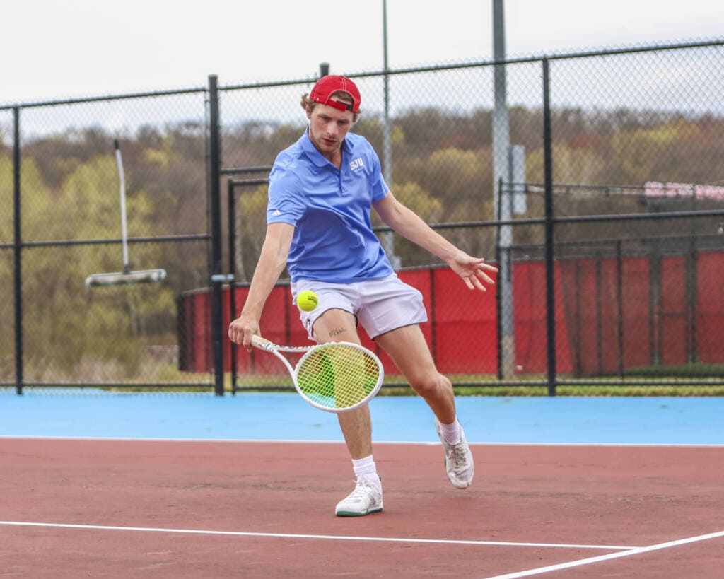 A tennis player in a blue shirt, white shorts, and a red cap hits a tennis ball with a racket mid-court on an outdoor court, with a chain-link fence and trees in the background.