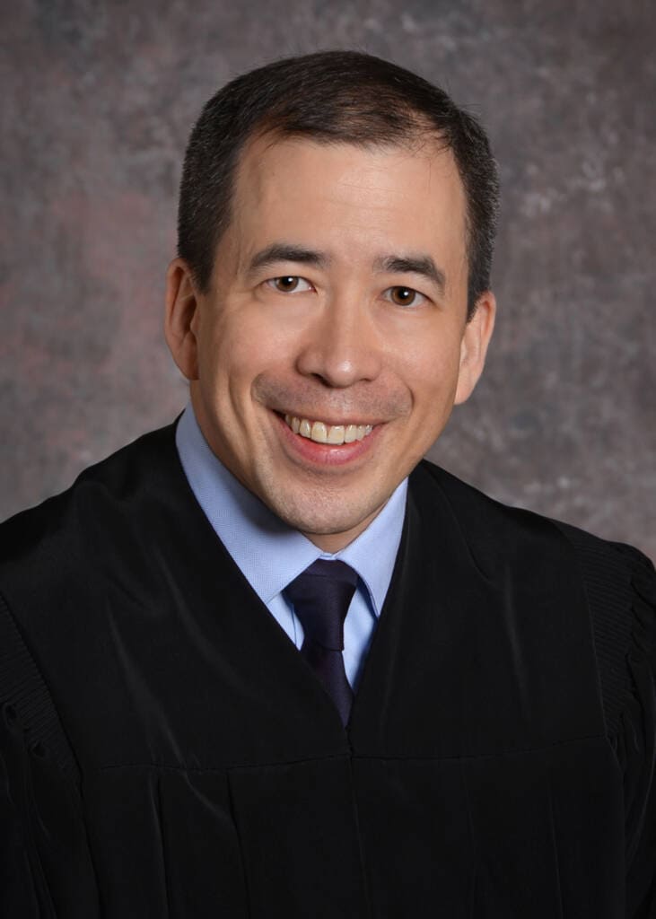 A man wearing a black judge's robe, light blue shirt, and dark tie smiles at the camera against a textured gray background.