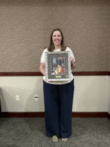 A woman stands indoors, smiling and holding a framed picture with a decorative border. The background is a plain, beige wall with a wooden trim and a patterned carpet.