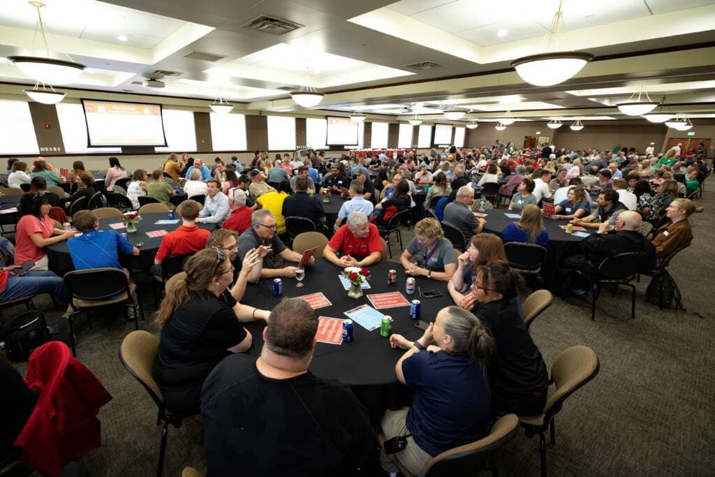 A large group of people sit at round tables in a spacious, well-lit conference room, engaged in conversations. Projector screens display information at the front, and canned drinks are visible on tables.