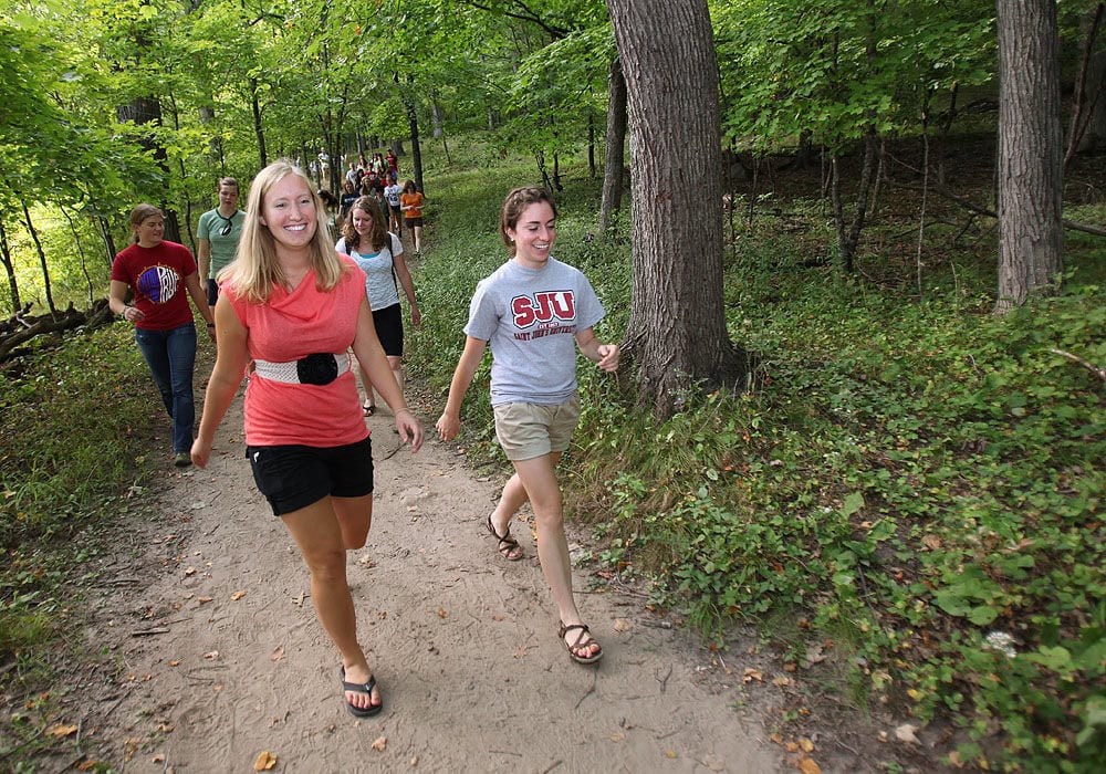 A group of people walk along a dirt trail in a green, wooded area. Two women in shorts and t-shirts lead the group, smiling as they enjoy the nature walk under tall trees.