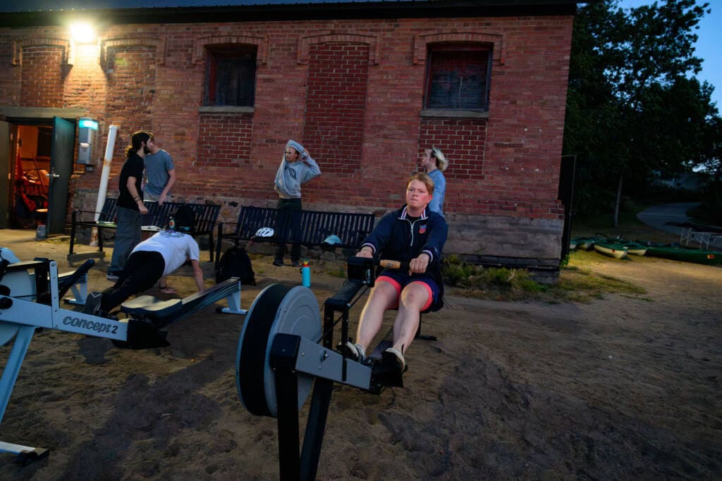 Several people exercise outside a brick building in the evening; one person uses a rowing machine in the foreground while others do push-ups or stretch near a bench.