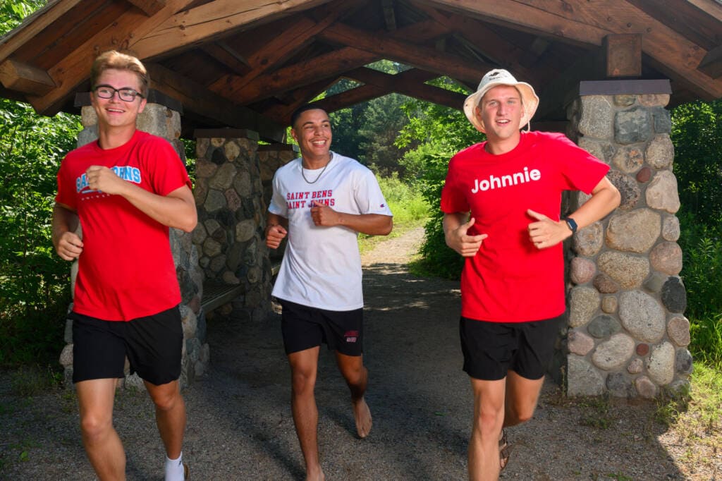 Three young men wearing athletic gear jog together on a gravel path under a wooden shelter. They are smiling and enjoying the outdoors, surrounded by greenery and sunlight.