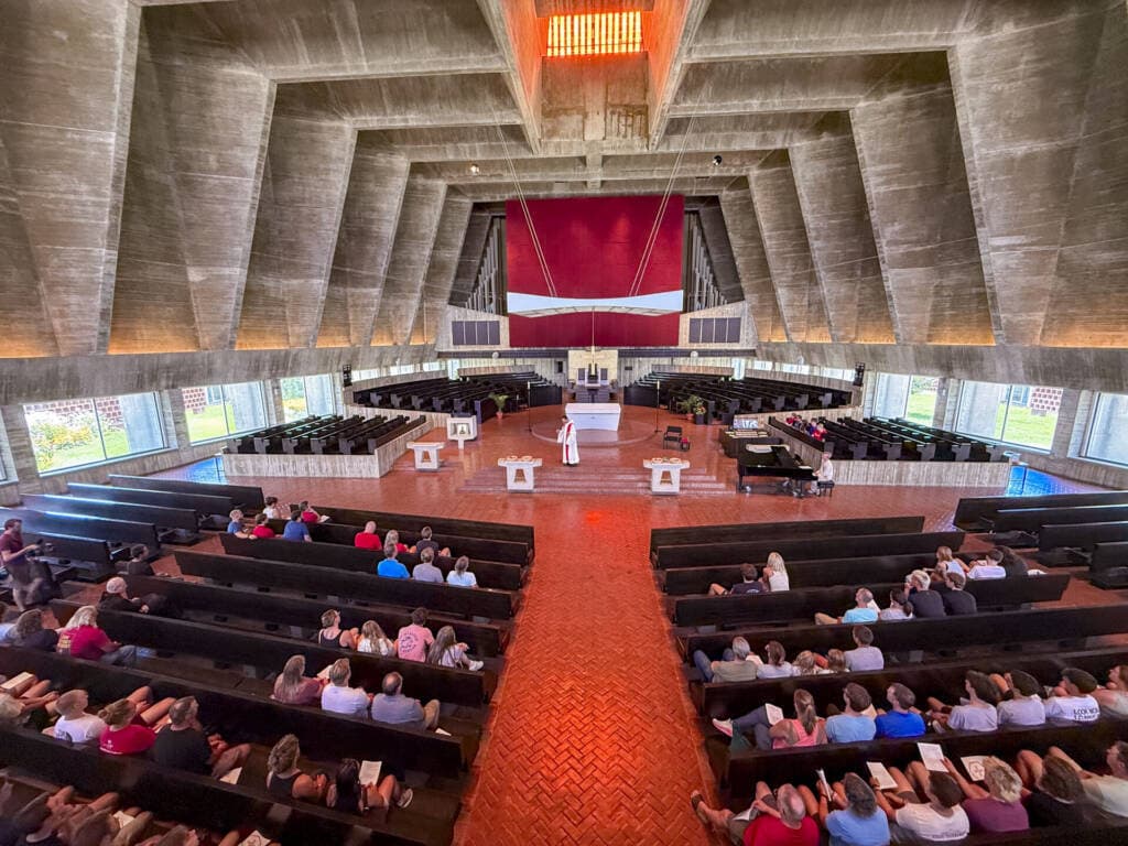 A wide-angle view of a modern church interior with geometric concrete ceilings, red brick floor, and large windows. A person stands at the altar while people sit in dark pews, facing forward and listening.