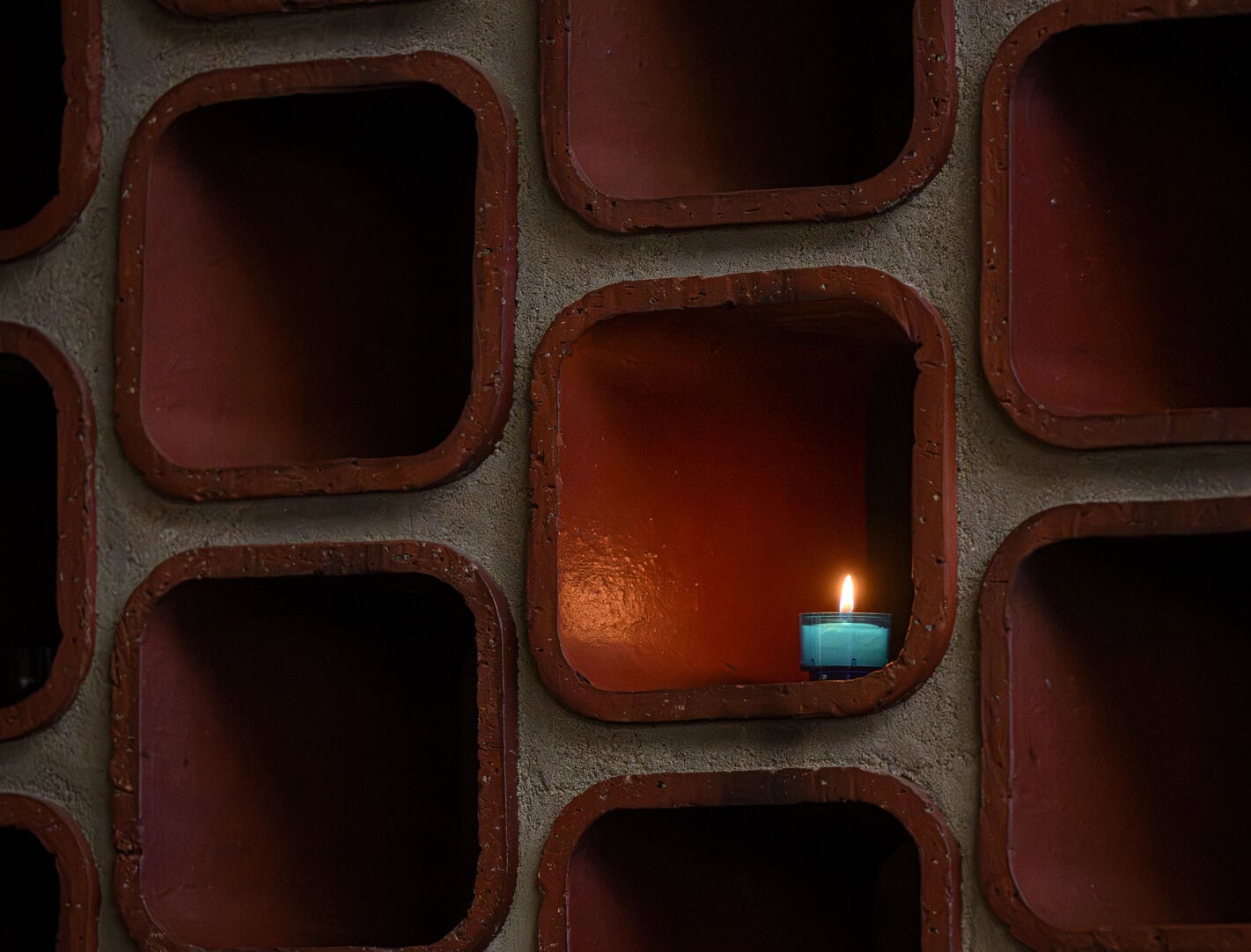 A blue tealight candle burns inside a single square cubby of a brown, grid-like wall with multiple empty cubbyholes, creating a warm, cozy contrast.