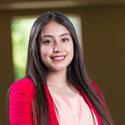 A young woman with long brown hair smiles at the camera. She is wearing a pink blouse and a red blazer. The background is softly blurred, featuring a large window with greenery outside.