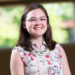 A young person with shoulder-length dark hair, wearing glasses and a sleeveless floral blouse, smiles warmly at the camera. They are also wearing a statement necklace. The background is softly blurred, suggesting a natural, well-lit setting.